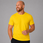 A smiling bald man with a beard wearing a bright yellow crew-neck t-shirt and black pants, standing against a solid gray studio background.