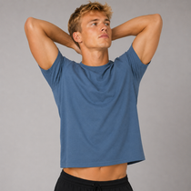 A man wearing a cropped, short-sleeve crewneck t-shirt in a muted slate blue, with his hands behind his head, posing against a solid grey studio background.