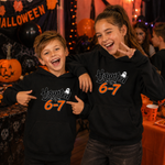 Two children smiling and pointing at matching black hoodies with a Halloween-themed graphic that reads 'Haunted by 6-7' in white and orange text, set in a decorated party environment with pumpkins and orange tablecloths.