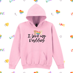 A light pink pullover hoodie with a kangaroo pocket and a graphic on the chest showing two bears holding a rainbow heart with the text 'I love my Daddies' in black script. The hoodie is displayed on a white background patterned with small rainbow hearts and the word 'LOVE'.