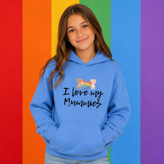 A young girl with long brown hair wearing a light blue hoodie with the text 'I love my Mummies' and a small cartoon bear graphic, standing in front of a vibrant rainbow-striped background.