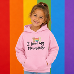 A young girl with her hair in a ponytail smiles while wearing a light pink hoodie with the text 'I love my Mummies' and a small rainbow heart graphic. She is standing in front of a vibrant rainbow-striped background.