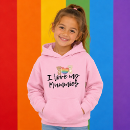 A young girl with her hair in a ponytail smiles while wearing a light pink hoodie with the text 'I love my Mummies' and a small rainbow heart graphic. She is standing in front of a vibrant rainbow-striped background.