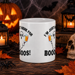 A white ceramic mug with a Halloween graphic design, displayed on a dark wooden table surrounded by spooky props including a jack-o'-lantern, lanterns, a skull, and autumn leaves.