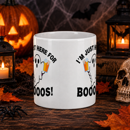 A white ceramic mug with a Halloween graphic design, displayed on a dark wooden table surrounded by spooky props including a jack-o'-lantern, lanterns, a skull, and autumn leaves.