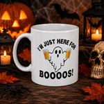A white ceramic mug with a Halloween graphic that reads 'I'M JUST HERE FOR BOOOOOS!' featuring a ghost holding two beer mugs. The mug is set on a dark wooden table surrounded by spooky props including a skull, a black spider, orange autumn leaves, a jack-o'-lantern, and lanterns with candles.