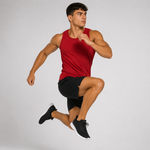 A man in a red sleeveless athletic tank top and black athletic shorts is captured mid-jump against a plain light gray studio background.