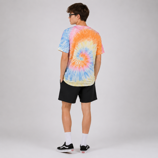 A model shown from the back wearing a vibrant tie-dye t-shirt with a spiral pattern in orange, yellow, blue, and pink, paired with black shorts, white socks, and black sneakers, set against a plain light gray studio background.