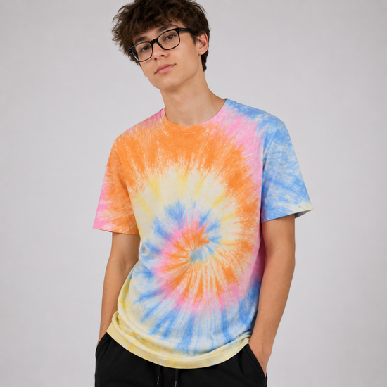 A young man with glasses wearing a vibrant tie-dye t-shirt with a spiral pattern of orange, pink, blue, and yellow, paired with black pants, against a plain light gray background.