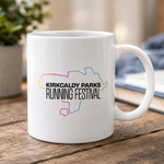A white ceramic mug with a colourful outline map of Kirkcaldy Park and the text 'KIRKCALDY PARKS RUNNING FESTIVAL' printed on the front, sitting on a wooden table in a blurred indoor setting.