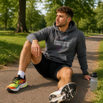 A man sitting on a paved path in a park wearing a dark Gray hoodie with 'KIRKGALEY PARKS RUNNING FESTIVAL' printed on the chest, black athletic shorts, and colourful running shoes, with a water bottle beside him.
