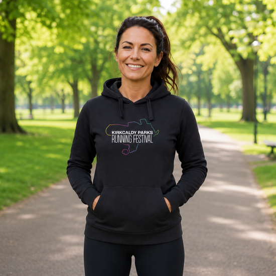 A woman stands on a paved path in a park wearing a black hoodie with a colourful map outline and the text 'Kirkcaldy Parks Running Festival' printed on the chest.