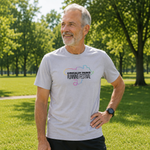 A smiling older man standing outdoors on a grassy park lawn wearing a light grey t-shirt with a colourful running map graphic and the text 'Kirkcaldy Parks Running Festival' on the chest.