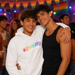 Two young men smiling and posing together at a party. The man on the left wears a white hoodie with the rainbow-colored text '#LOVEWINS!' across the chest, and the man on the right wears a black tank top.