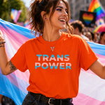 A woman at a pride parade wearing a bright orange t-shirt with 'TRANS POWER' printed in white and light blue letters, holding a pink, blue, and white transgender pride flag.