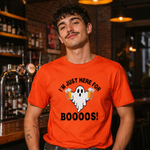 A man wearing a bright orange graphic t-shirt in a dimly lit bar setting. The shirt features a white cartoon ghost holding two beer mugs with the text 'I'M JUST HERE FOR BOOOOS!' printed in black.