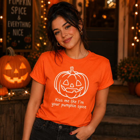 A woman wearing a bright orange graphic t-shirt with a white pumpkin illustration and the text 'Kiss me like I'm your pumpkin spice'. She is posing in front of a Halloween-themed storefront with pumpkins and warm string lights.