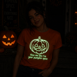 A woman wearing a glowing orange t-shirt with a green pumpkin graphic and the text 'Kiss me like I'm your pumpkin spice' in a dark, Halloween-themed setting with a lit jack-o'-lantern.