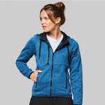 A woman wearing a heathered blue zip-up hoodie with black trim, black drawstrings, and a black inner hood lining, photographed against a solid grey studio background.