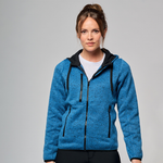 A woman wearing a heathered blue zip-up hooded jacket with black trim, zippers, and drawstrings, layered over a white t-shirt and black pants, against a solid grey studio background.