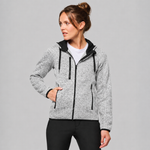 A woman wearing a light grey heather zip-up hoodie with black trim, black drawstrings, and black side zippers, paired with black leggings, against a solid grey studio background.