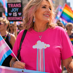 A woman at a pride parade wearing a bright pink t-shirt with a transgender pride cloud graphic. She is smiling and holding a trans flag, with other participants and rainbow flags blurred in the background.