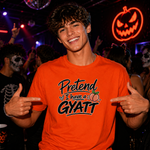 A smiling young man at a nightclub or party wearing a bright orange graphic T-shirt that reads 'Pretend I have a GYATT' with a peach illustration. The background is dark with red neon Halloween decorations and other partygoers.