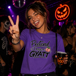 A woman at a Halloween party wearing a purple graphic t-shirt that reads 'Pretend I have a gyatt' with a peach graphic, holding a black 'BOO' cup and making a peace sign.