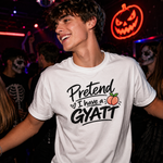 A smiling young man in a dimly lit club wearing a white graphic T-shirt that reads 'Pretend I have a GYATT' with a small peach illustration. Other people and a glowing orange pumpkin sign are visible in the background.