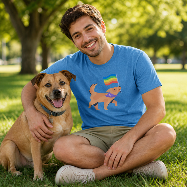A smiling man sitting on grass in a park with a tan dog beside him. The man is wearing a bright blue graphic T-shirt featuring a cartoon dog holding a rainbow flag and a small purple heart, along with khaki shorts and white sneakers.