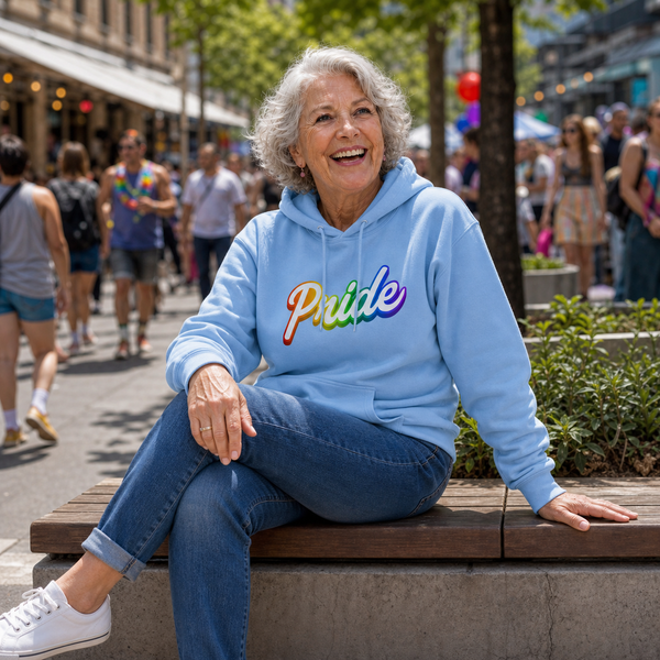 An older woman with grey hair sits smiling on a wooden bench in a crowded outdoor street setting. She is wearing a light blue hoodie with the word 'Pride' in rainbow lettering across the chest, blue jeans, and white sneakers.