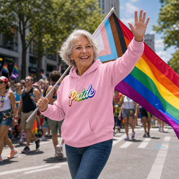 An older woman smiling and waving while marching in a pride parade, wearing a pink hoodie with the word 'Pride' in rainbow letters and carrying a large pride flag.