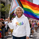 A smiling older woman at a pride parade holds a rainbow flag and wears a white hoodie with the word 'Pride' in rainbow lettering. The background shows a crowded outdoor street scene with other participants and buildings.