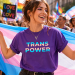 A woman at a pride parade wearing a purple t-shirt with 'TRANS POWER' printed in pink, white, and blue gradient letters. She is holding a pink, white, and blue transgender flag, with a crowd and rainbow flags blurred in the background.