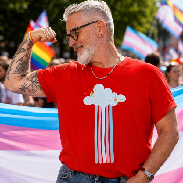A smiling man with gray hair and glasses flexes his arm in a pride setting, wearing a bright red t-shirt with a white cloud graphic that is splitting into pink and light blue rain-like streaks. Pride flags are visible in the blurred background.