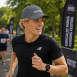 A smiling male runner in a park wearing a black New Balance t-shirt and a grey Kilkenny & Districts cap, with race banners in the background.