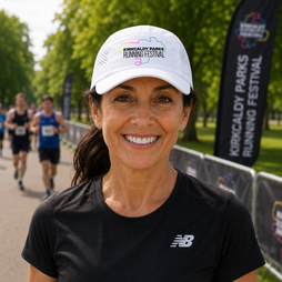 A smiling woman wearing a white Kirkcaldy Parks Running Festival cap and a black New Balance t-shirt, standing outdoors with race participants blurred in the background.