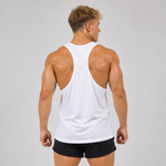 A muscular man viewed from the back wearing a white racerback tank top and black athletic shorts, standing against a light gray studio background.