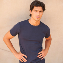 A man wearing a dark navy blue crewneck t-shirt, standing with hands on hips against a textured beige wall.