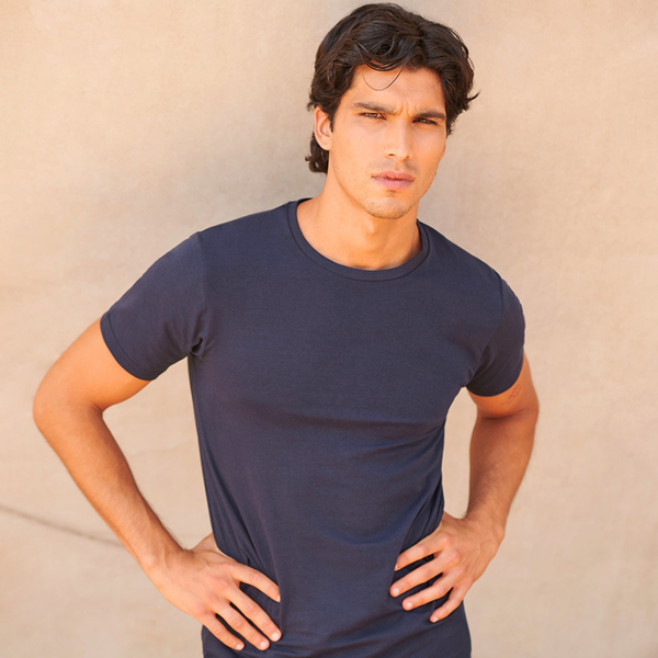 A man wearing a dark navy blue crewneck t-shirt, standing with hands on hips against a textured beige wall.