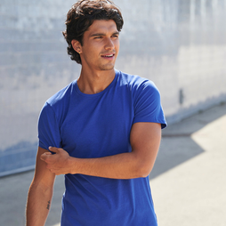 A man wearing a bright royal blue short-sleeve crewneck t-shirt, standing outdoors against a blurred concrete wall background.