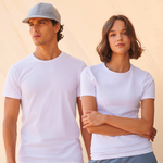Two models wearing plain white short-sleeve crewneck t-shirts, standing against a light beige background. The man on the left wears a light gray baseball cap, and the woman on the right has her arms crossed.