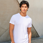 A man wearing a plain white short-sleeve crewneck t-shirt, standing against a textured beige concrete wall.