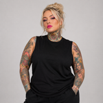 A plus-size model with tattoos wearing a black sleeveless mock-neck top, standing against a solid gray studio background.