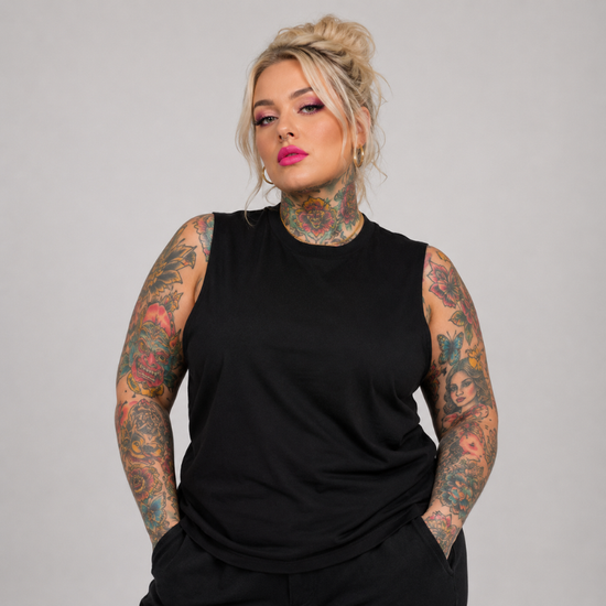 A plus-size model with tattoos wearing a black sleeveless mock-neck top, standing against a solid gray studio background.