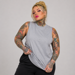 A plus-size model with tattoos wearing a light gray sleeveless mock-neck t-shirt and black pants, posing against a solid gray studio background.