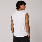 A man with dark hair and tattoos is shown from the back wearing a plain white sleeveless t-shirt and black pants, standing against a light gray studio background.