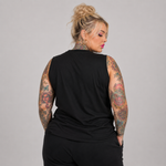 A woman with blonde hair in a bun and colorful arm tattoos, shown from the back wearing a black sleeveless top and black pants against a solid gray studio background.