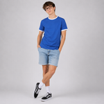 A young man wearing glasses, a bright blue ringer t-shirt with white trim, light blue denim shorts, white socks, and black Vans sneakers, standing against a light gray studio background.