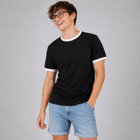 A man wearing a black short-sleeve ringer t-shirt with white trim on the collar and sleeve cuffs, paired with light blue denim shorts, standing against a solid gray studio background.